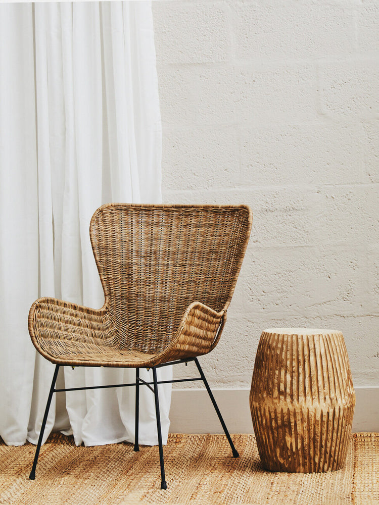 Curved natural rattan chair with a woven design, featuring black metal legs, positioned next to a wooden side table. The background includes a light-colored wall and white curtains, with a warm wooden floor visible