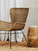 Curved design natural rattan chair with a woven backrest and armrests, set on black metal legs, next to a wooden side table, against a light-colored wall and white curtains, with a wooden floor