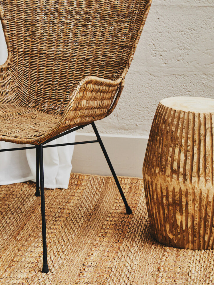 Curved design natural rattan chair with a woven seat and backrest, supported by black metal legs, positioned next to a round, textured wooden side table on a light-colored floor