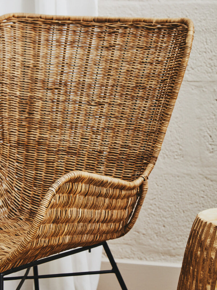 Curved natural rattan chair with a woven texture and a sleek black metal frame, showcasing a warm, earthy aesthetic. The chair's backrest features a distinctive shape that enhances its modern design, while a small side table made of natural materials is partially visible beside it