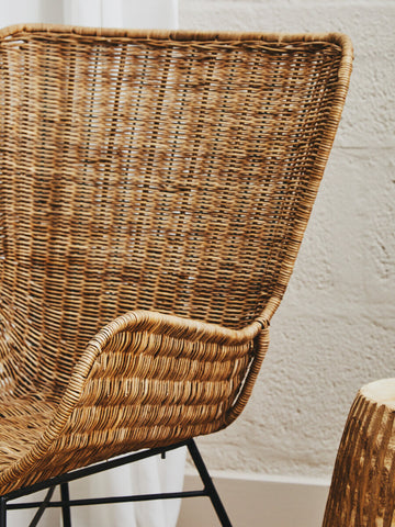 Curved natural rattan chair with a woven texture and a sleek black metal frame, showcasing a warm, earthy aesthetic. The chair's backrest features a distinctive shape that enhances its modern design, while a small side table made of natural materials is partially visible beside it