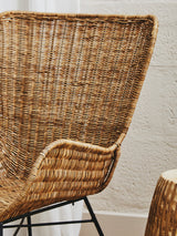 Curved natural rattan chair with a woven texture and a sleek black metal frame, showcasing a warm, earthy aesthetic. The chair's backrest features a distinctive shape that enhances its modern design, while a small side table made of natural materials is partially visible beside it