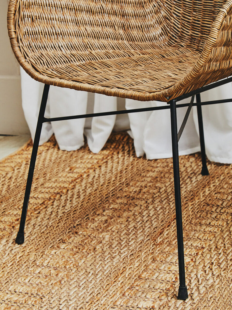 Curved natural rattan chair with a woven seat and backrest, supported by sleek black metal legs, placed on a textured natural fiber rug with a light-colored backdrop