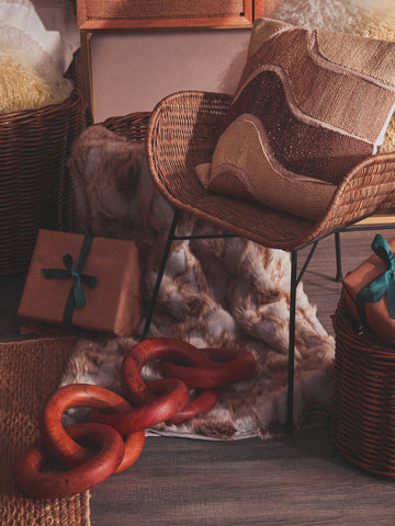 Curved design natural rattan chair with a light brown finish, surrounded by decorative hats, a red chain, and various textured materials, set against a warm, softly lit background