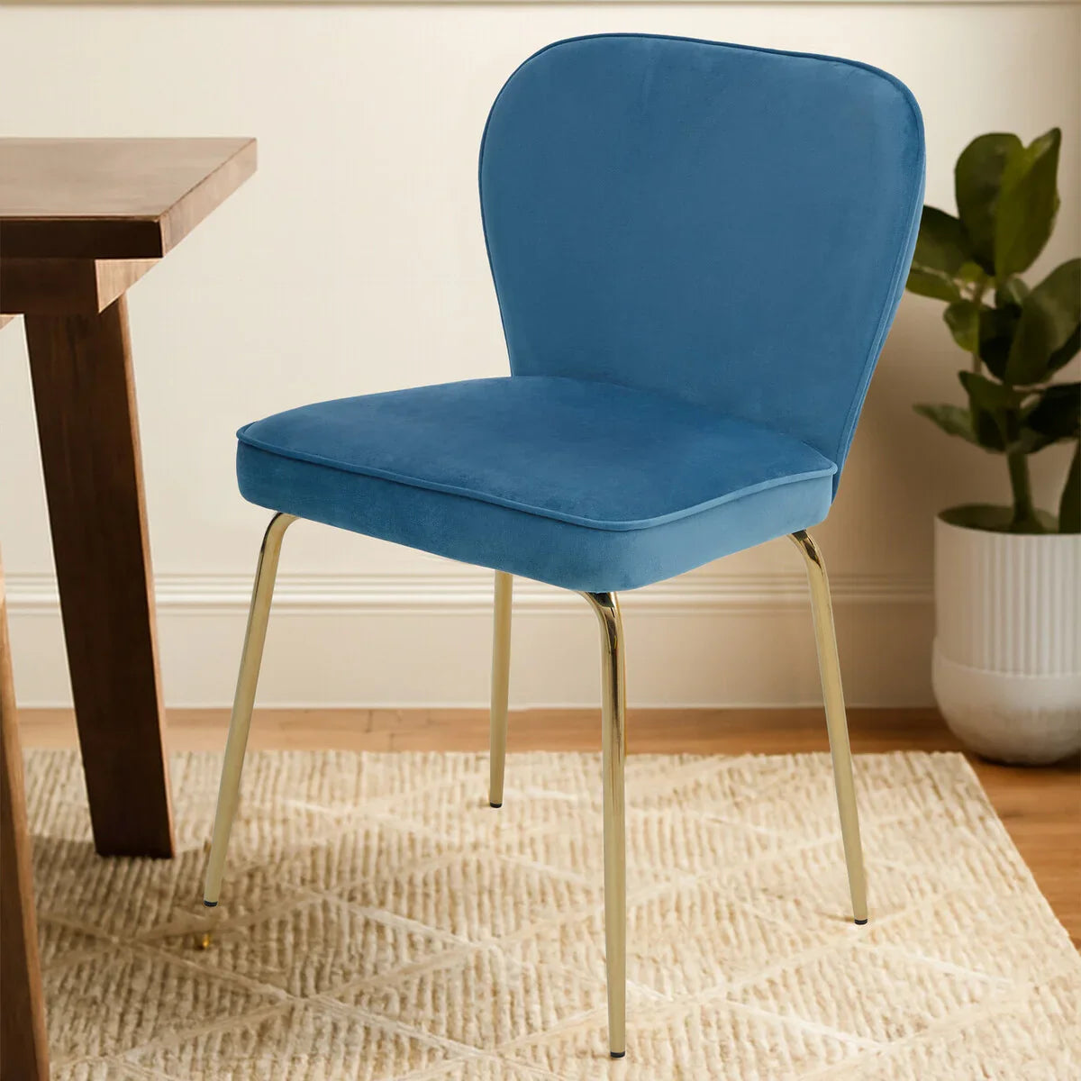 Blue velvet dining chair with gold metal legs beside wooden table on patterned rug
