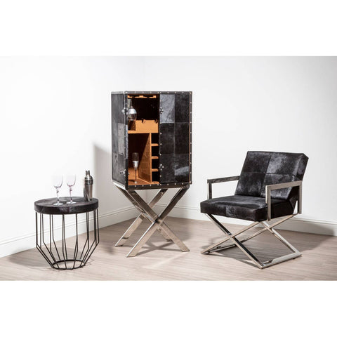 A stylish drinks cabinet with a hair-on-leather finish, featuring a cross base design, positioned next to a modern black leather chair and a round wire side table. The cabinet is open, revealing wooden shelves inside, set against a neutral wall and light-colored floor