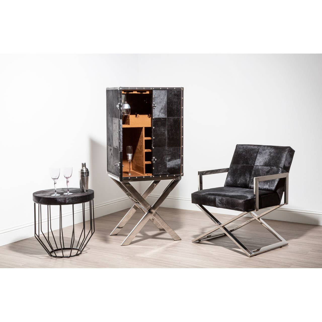 A stylish drinks cabinet with a hair-on-leather finish, featuring a cross base design, positioned next to a modern black leather chair and a round wire side table. The cabinet is open, revealing wooden shelves inside, set against a neutral wall and light-colored floor