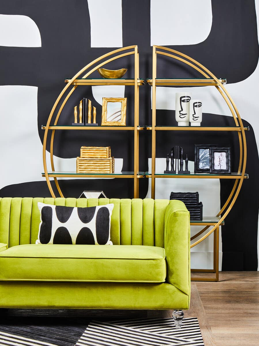 A vibrant green velvet three-seat sofa with vertical tufting, positioned in a modern living space. Behind the sofa, a circular gold shelving unit displays decorative items, including books and vases, against a bold black and white wall. A black and white patterned pillow rests on the sofa, complementing the overall contemporary aesthetic. The floor features light wood planks, adding warmth to the scene