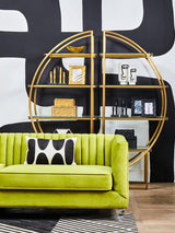A vibrant green velvet three-seat sofa with vertical tufting, positioned in a modern living space. Behind the sofa, a circular gold shelving unit displays decorative items, including books and vases, against a bold black and white wall. A black and white patterned pillow rests on the sofa, complementing the overall contemporary aesthetic. The floor features light wood planks, adding warmth to the scene