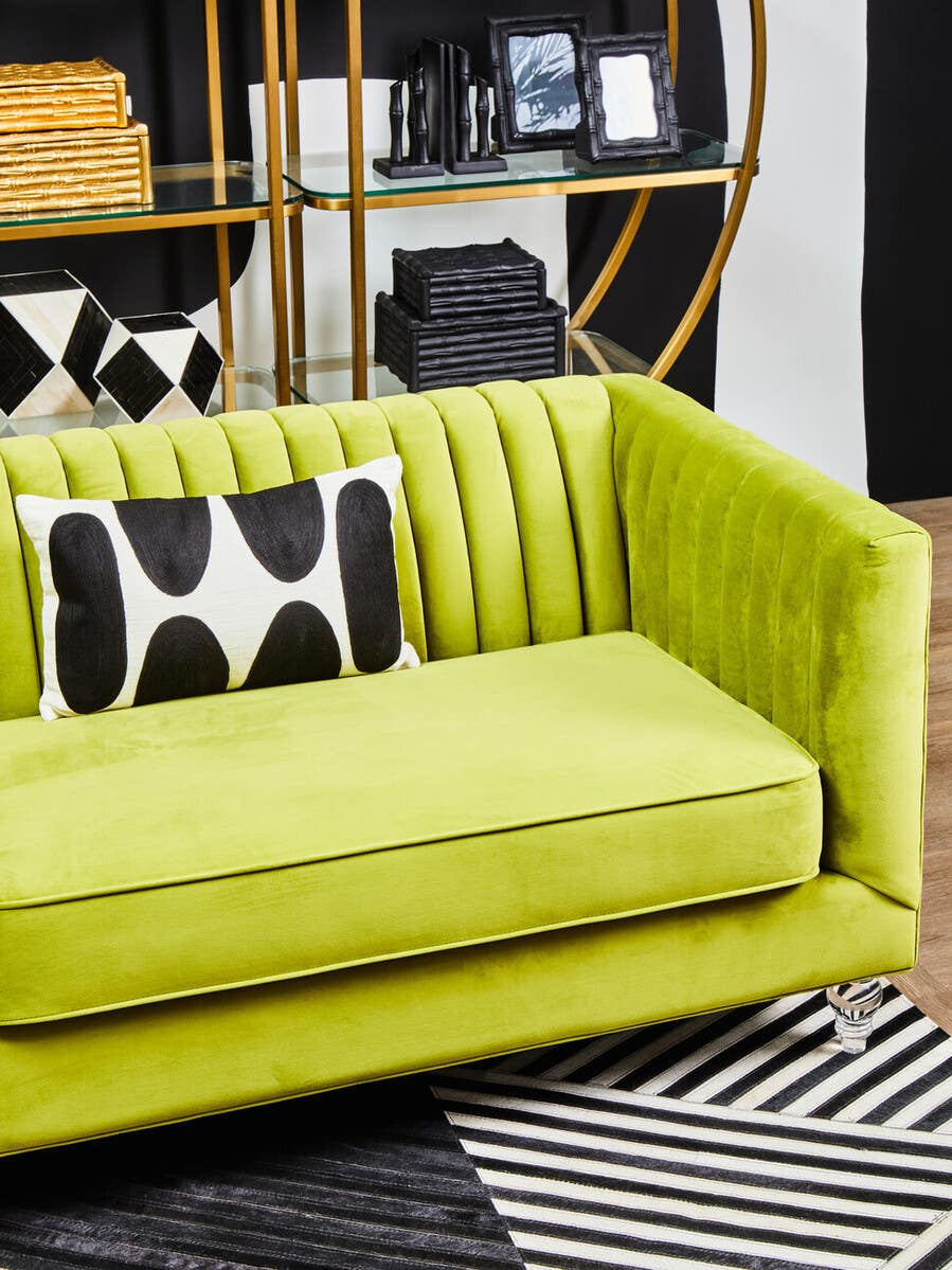 A vibrant green velvet sofa with a modern design featuring a three-seat configuration. The sofa has a textured backrest with vertical pleats and a plush seat cushion. A black and white patterned throw pillow is placed on one side. In the background, there is a stylish shelving unit with decorative items and a contrasting black and white rug underneath