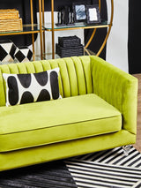 A vibrant green velvet sofa with a modern design featuring a three-seat configuration. The sofa has a textured backrest with vertical pleats and a plush seat cushion. A black and white patterned throw pillow is placed on one side. In the background, there is a stylish shelving unit with decorative items and a contrasting black and white rug underneath
