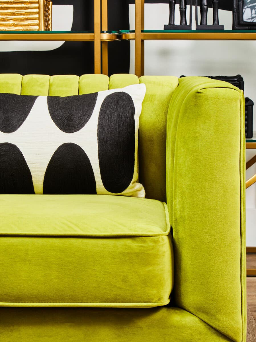 A close-up view of a green velvet sofa featuring a modern design, with a plush seat and armrest. The sofa includes a decorative black and white patterned pillow, set against a backdrop of wooden shelves displaying various decorative items