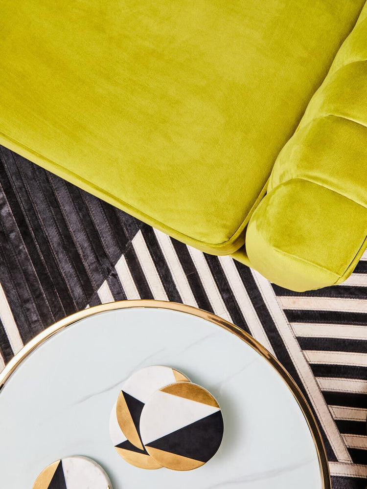 A close-up view of a vibrant green velvet fabric, showcasing its soft texture, alongside a round plate with a geometric design in black, white, and gold. The background features a striped pattern in black and white, adding contrast to the scene
