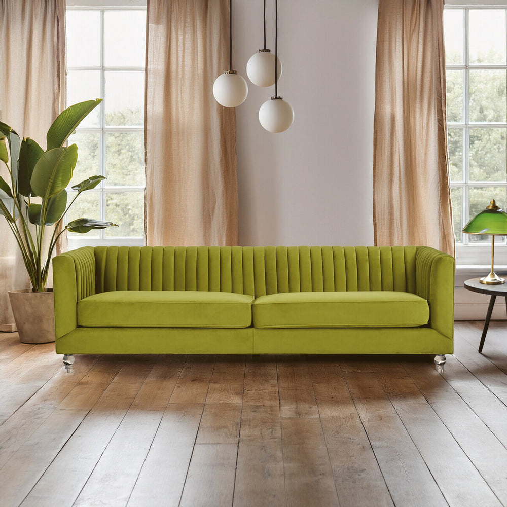 A vibrant green velvet sofa with a modern design, featuring a ribbed texture and three spacious seats. The sofa is positioned in a bright room with large windows, allowing natural light to illuminate the space. Soft curtains frame the windows, and a potted plant adds a touch of greenery to the setting. Three pendant lights hang above the sofa, enhancing the contemporary aesthetic. The floor is made of warm wooden