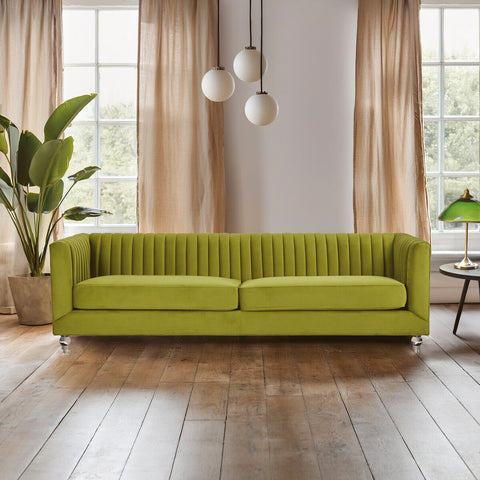 A vibrant green velvet sofa with a modern design, featuring a ribbed texture and three spacious seats. The sofa is positioned in a bright room with large windows, allowing natural light to illuminate the space. Soft curtains frame the windows, and a potted plant adds a touch of greenery to the setting. Three pendant lights hang above the sofa, enhancing the contemporary aesthetic. The floor is made of warm wooden