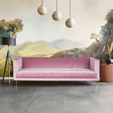 A three-seat pink sofa with a sleek, modern design featuring a smooth fabric upholstery and minimalist wooden legs. The sofa is positioned against a mural depicting rolling hills and trees, with three round pendant lights hanging above. A tall plant in a pot is visible to the right, complementing the contemporary decor