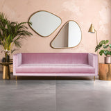 A stylish three-seat sofa in a soft pink fabric, featuring a modern design with clean lines and a low back. The sofa is positioned against a light pink wall and is complemented by two decorative mirrors above it. A potted plant and a small side table with a lamp are visible to the left, enhancing the contemporary living space. The flooring is a smooth gray surface