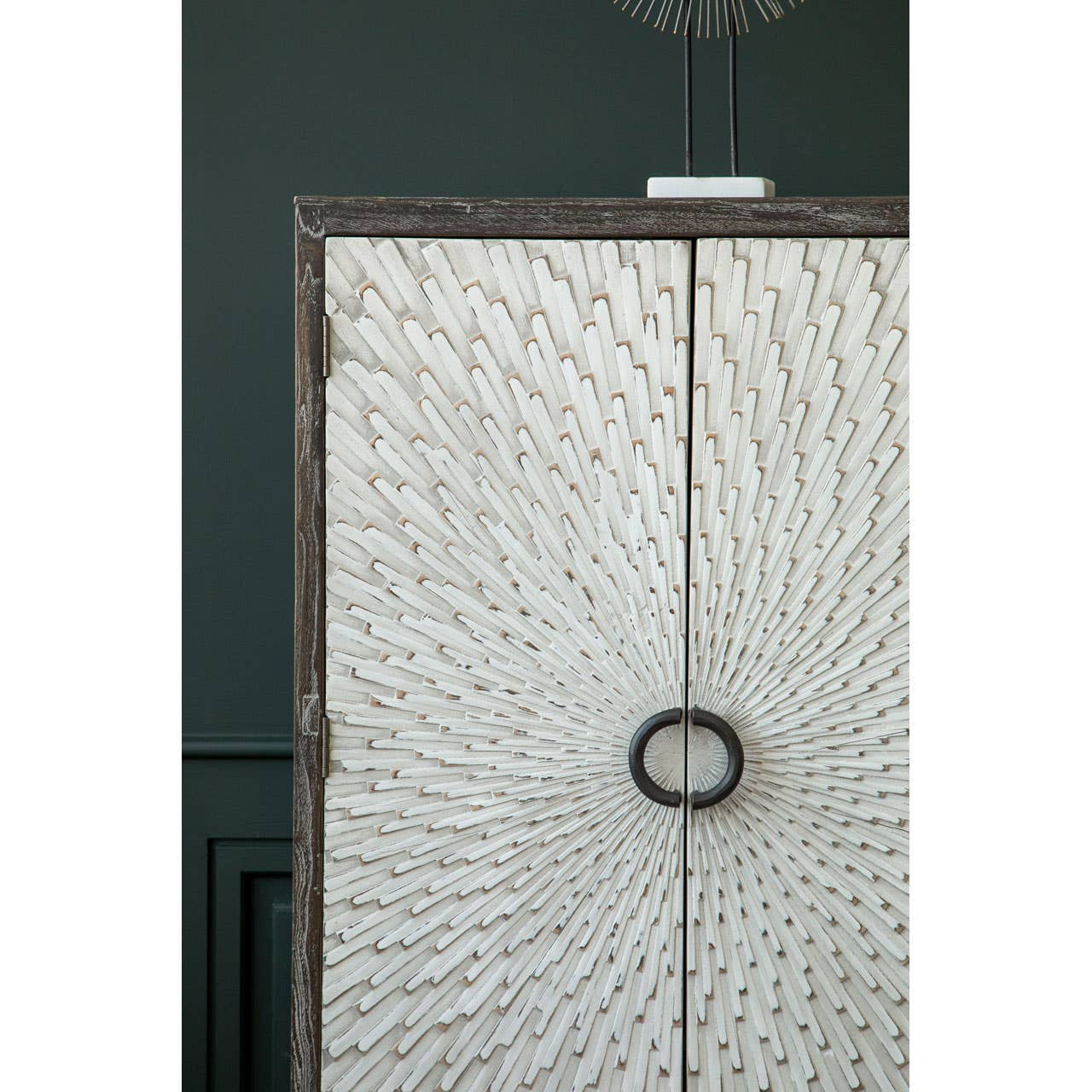 Milan Two Door Elm Wood Wine Cabinet featuring a textured front with a circular handle, set against a dark green wall. The cabinet showcases a natural wood finish with intricate patterns, emphasizing its elegant design