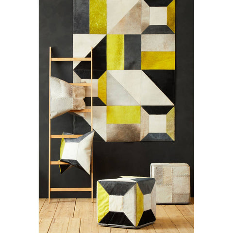 A geometric design pouffe featuring a mix of hair-on leather in shades of yellow, black, and cream. The pouffe is placed on a wooden floor, with a backdrop of a wall displaying a similar geometric pattern in various colors. A wooden ladder leans against the wall, adding to the modern decor aesthetic