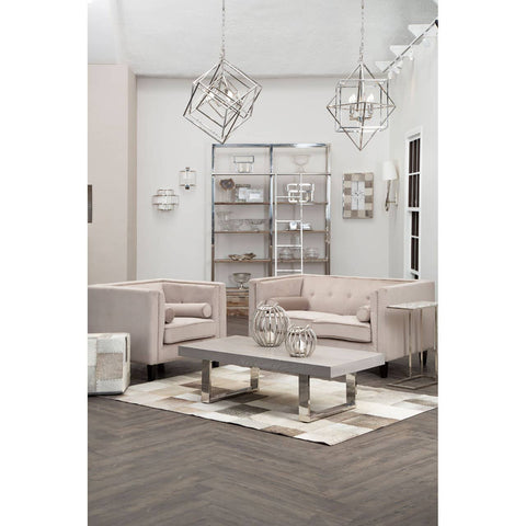 A stylish grey pouffe placed in a modern living room setting, surrounded by light-colored sofas and a coffee table. The room features a neutral color palette with decorative wall art and contemporary light fixtures hanging from the ceiling. A soft area rug is positioned under the coffee table, enhancing the cozy atmosphere
