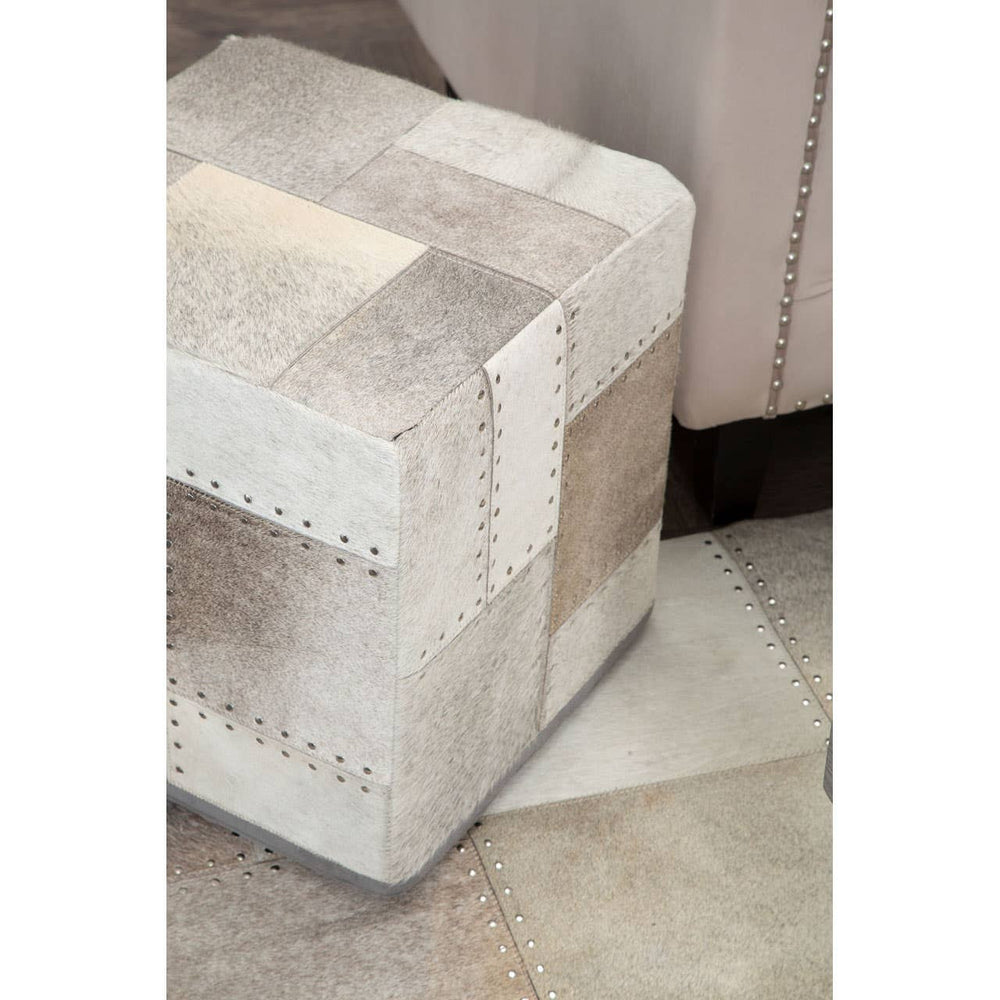 Square pouffe in a soft grey color with a textured patchwork design, featuring light and dark shades of grey and beige. The pouffe is placed on a patterned floor, partially visible in the image, with a neutral-toned wall in the background