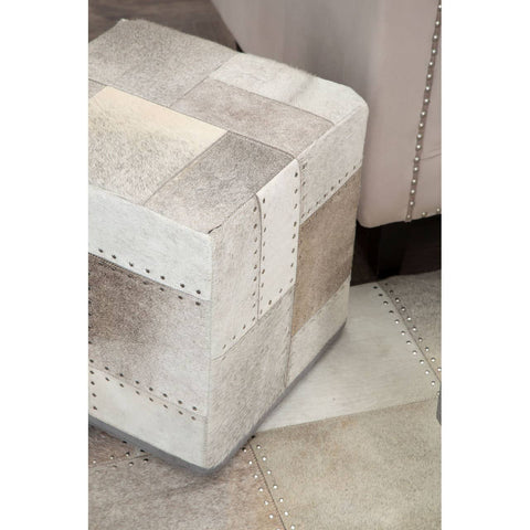 Square pouffe in a soft grey color with a textured patchwork design, featuring light and dark shades of grey and beige. The pouffe is placed on a patterned floor, partially visible in the image, with a neutral-toned wall in the background