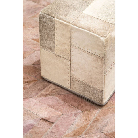 A square pouffe with a patchwork design in various shades of grey, featuring a smooth top and a sturdy base, placed on a wooden floor