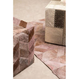 A grey pouffe with a textured surface, placed next to a light-colored cube-shaped pouffe, on a patterned rug featuring soft pink and beige tones. The background is a neutral, light-colored floor