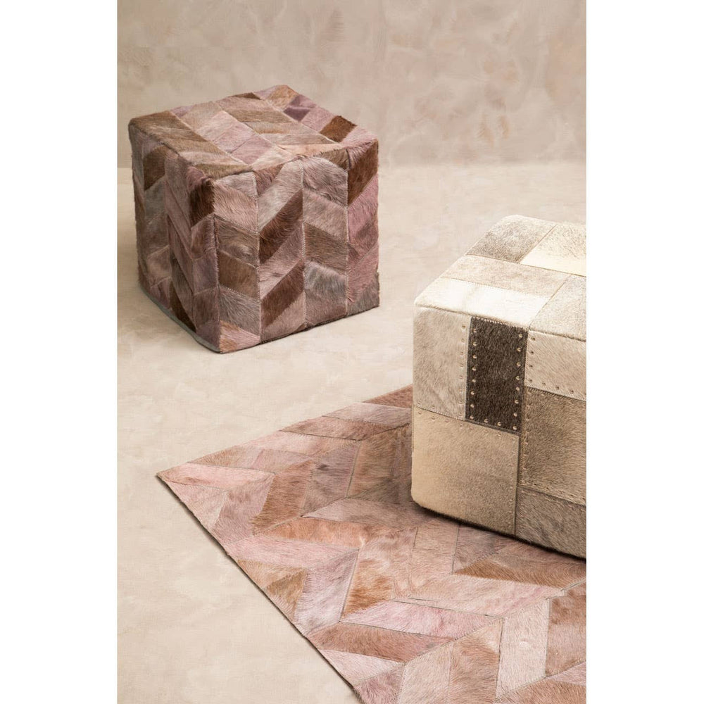 A textured grey pouffe with a geometric design sits on a light-colored floor, accompanied by a similarly styled pouffe in a warm, earthy tone. The background features a smooth, neutral wall that complements the furniture