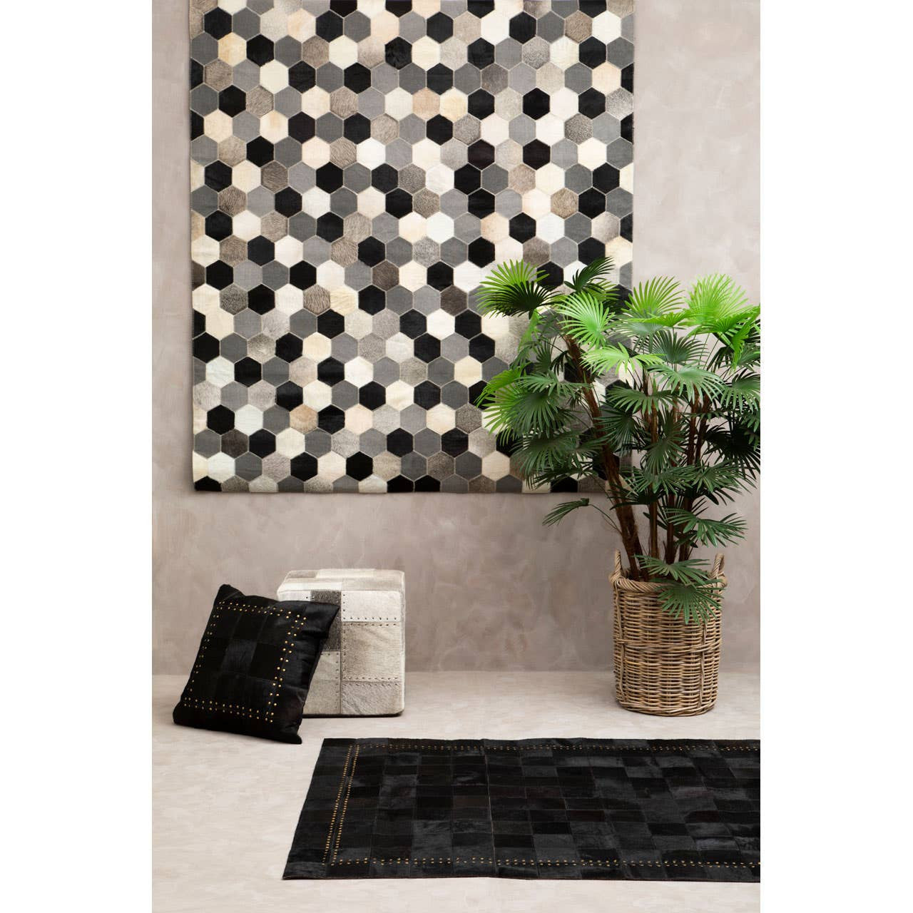 A grey pouffe placed in a stylish living space, accompanied by a patterned wall art piece in black and white, a potted plant, a decorative pillow, and a textured black rug on the floor