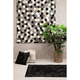 A grey pouffe placed in a stylish living space, accompanied by a patterned wall art piece in black and white, a potted plant, a decorative pillow, and a textured black rug on the floor