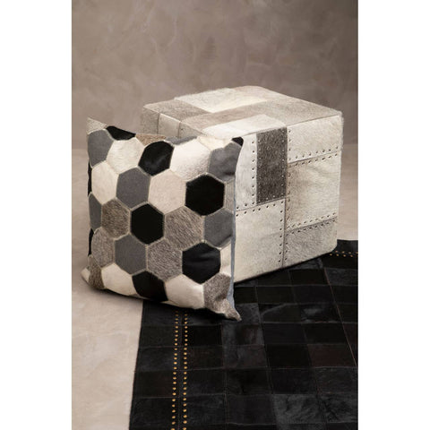 A grey pouffe with a textured surface, featuring a geometric design in shades of grey and black, sits on a dark rug. The pouffe has a cube shape and is adorned with hexagonal patterns, adding a modern touch to the decor