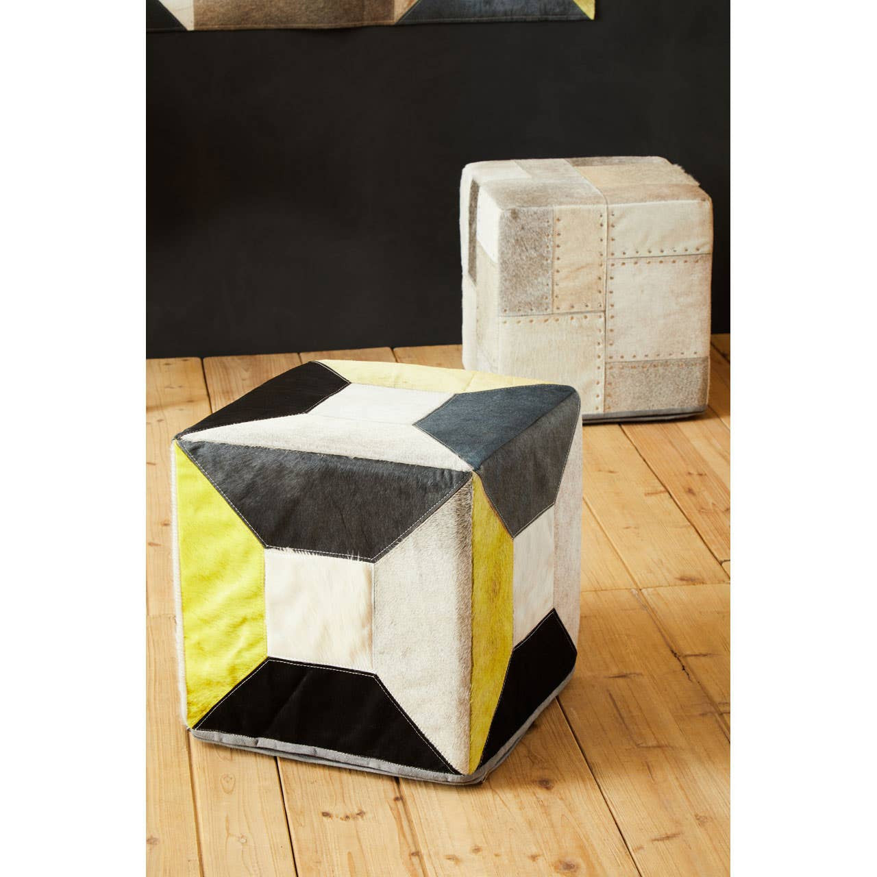 A geometric pouffe featuring a combination of grey, black, and yellow panels, with a textured surface, placed on a wooden floor. In the background, a similar pouffe is partially visible against a dark wall