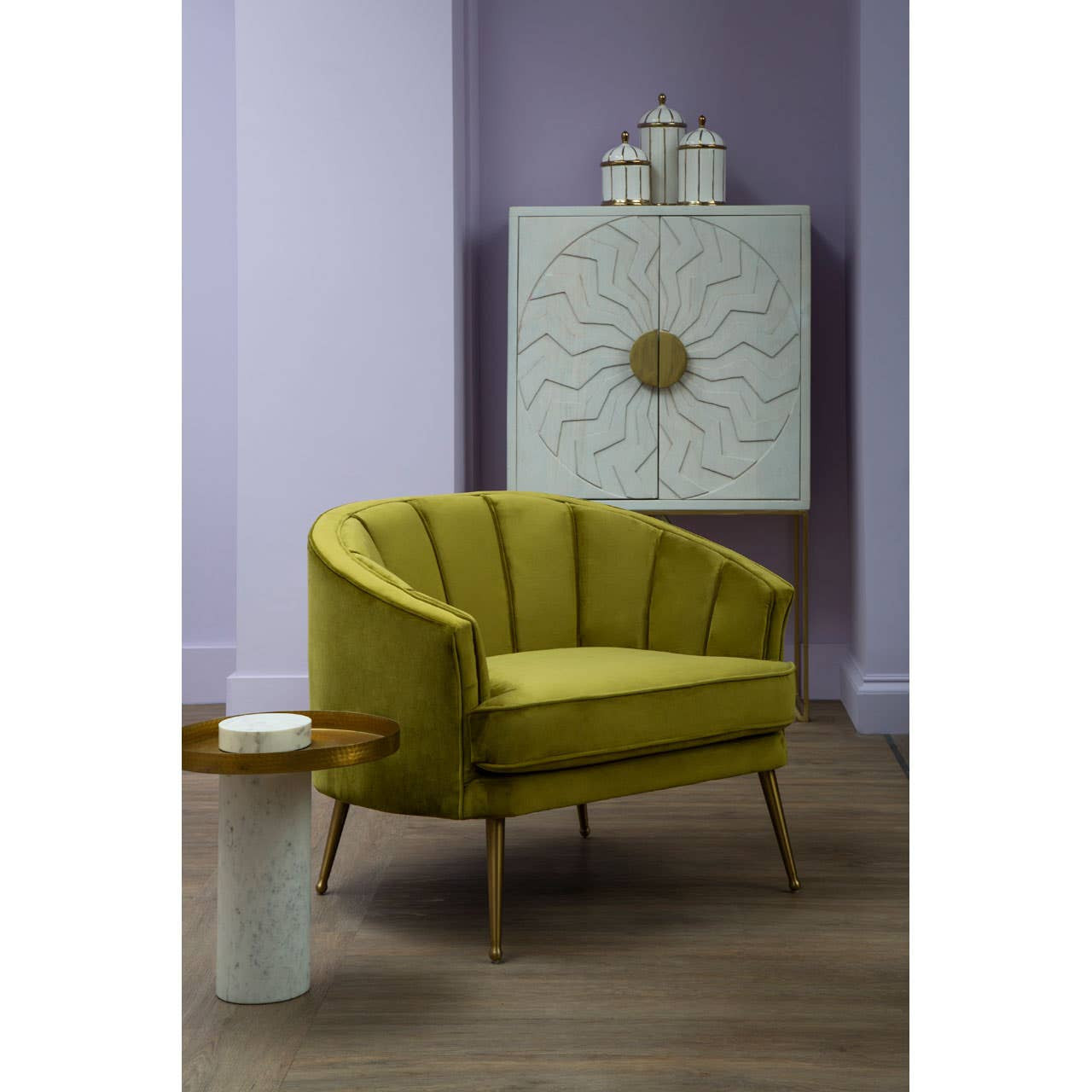 A stylish yellow accent chair with a curved backrest and wooden legs is positioned next to a small round side table. Behind the chair, a white wash mango wood cabinet features a textured front with a circular handle, set against a soft purple wall. The floor is a light wood finish, adding warmth to the scene