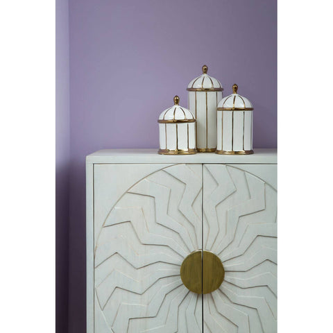 White wash mango wood cabinet featuring a sunburst design on the front, with two doors and a round brass handle. On top of the cabinet, three decorative jars with white and gold accents are arranged against a soft purple wall