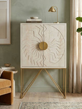 White wash mango wood cabinet with a textured circular design on the front, featuring a round brass handle. The cabinet stands on slender, angled gold legs and is positioned against a soft green wall. A small decorative lamp and a stack of books are placed on top, with a cozy beige armchair visible to the left