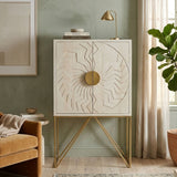 White wash mango wood cabinet with a textured front featuring a circular design and a round gold handle, supported by slender gold legs. The cabinet is placed against a green wall, with a cozy beige sofa and a potted plant visible in the background. Natural light filters through a nearby window, enhancing the warm tones of the room