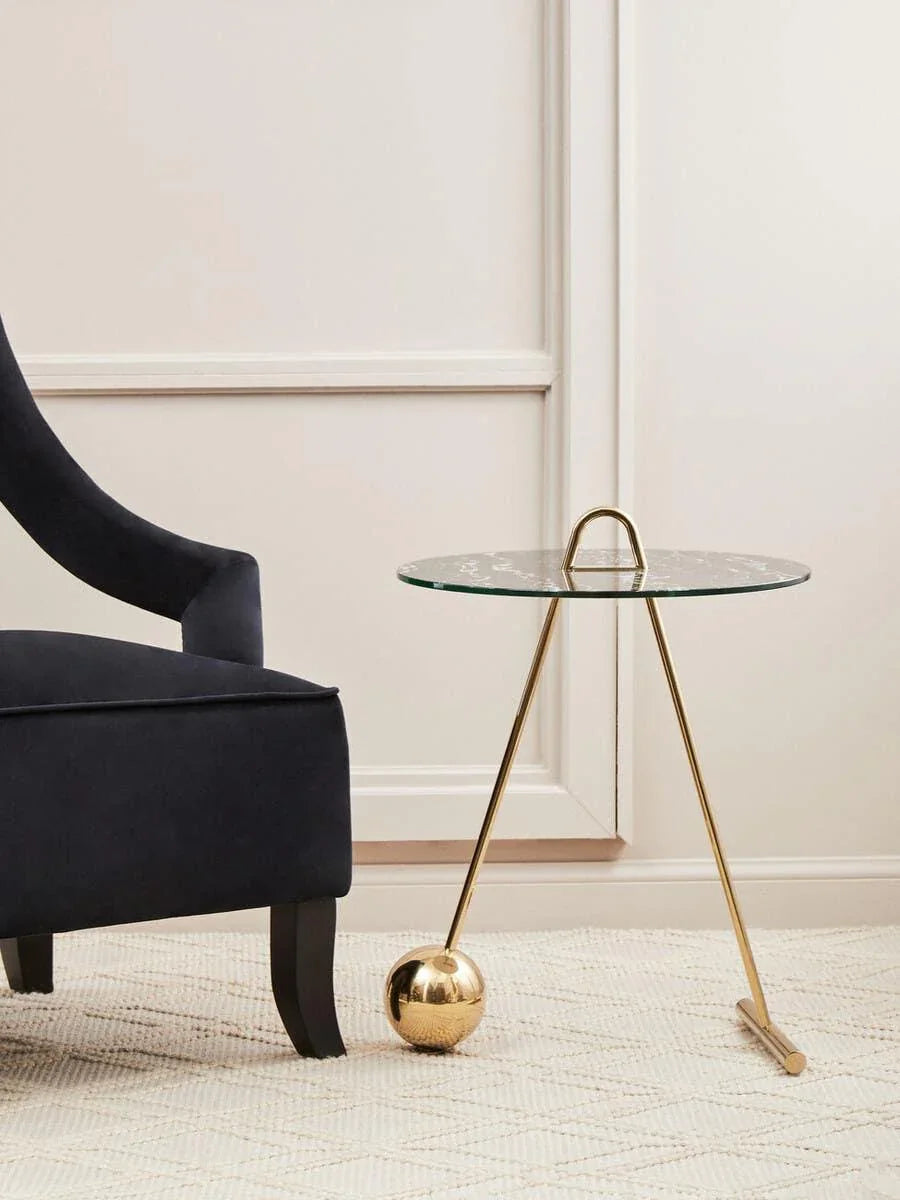Glass side table with gold metal base beside a navy blue armchair on textured rug