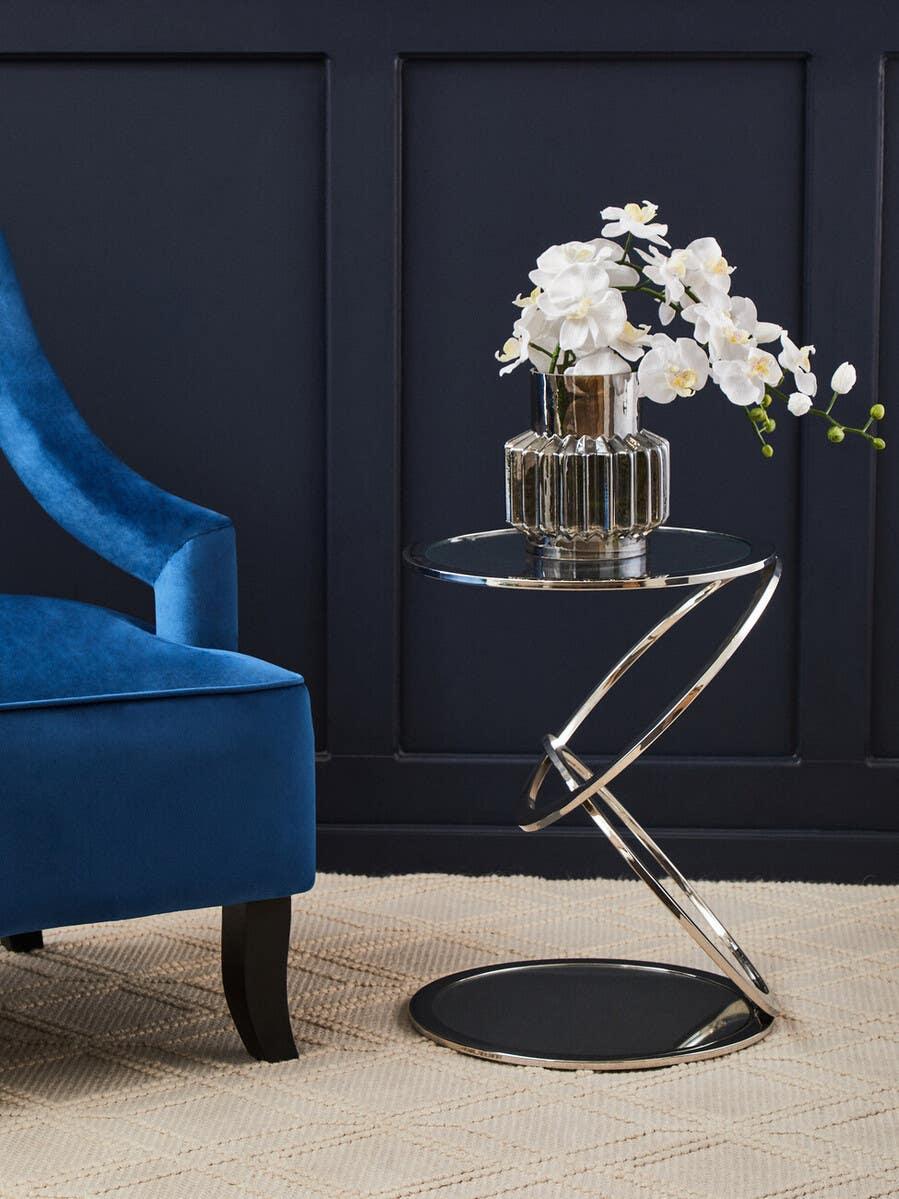 Modern chrome side table with round glass top, vase of white flowers, beside blue velvet armchair