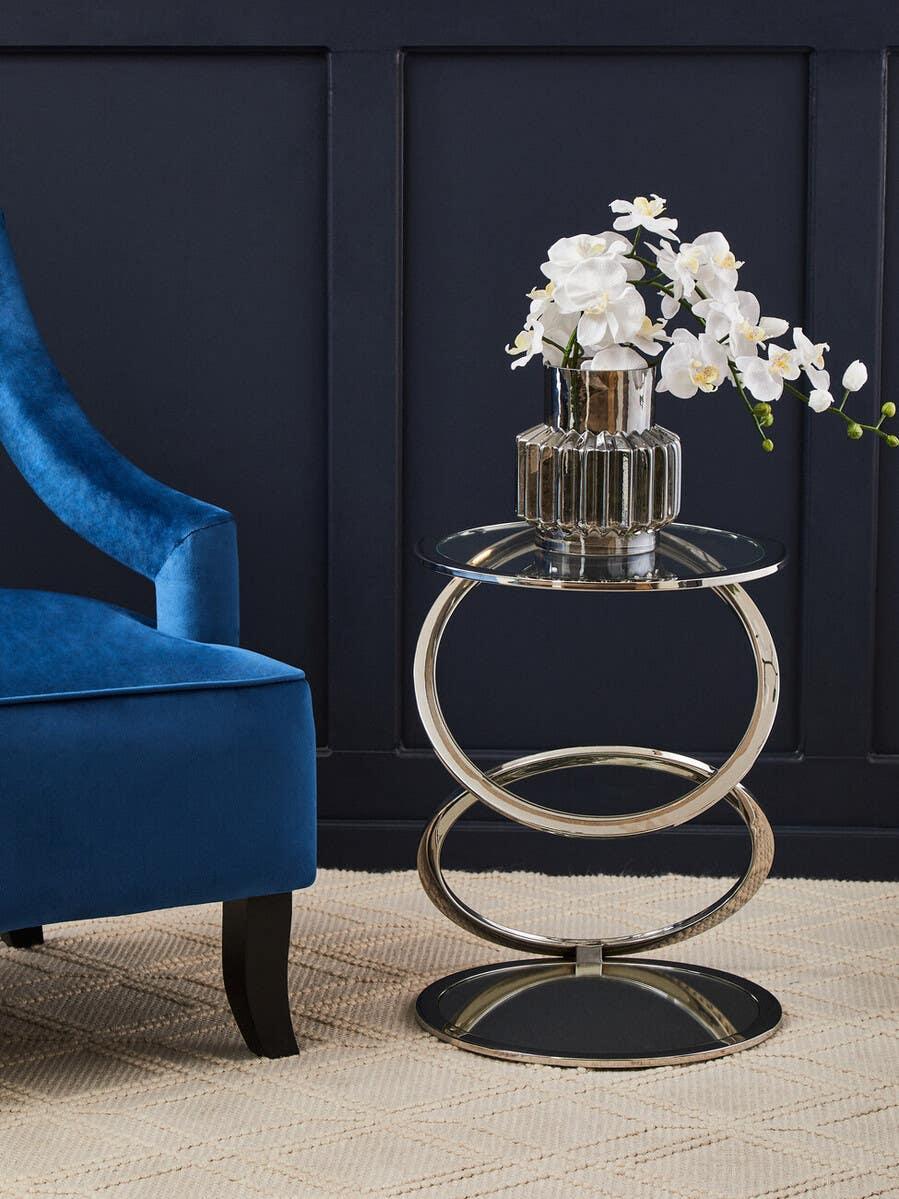 Glass top side table with chrome base and orchid vase next to blue velvet armchair
