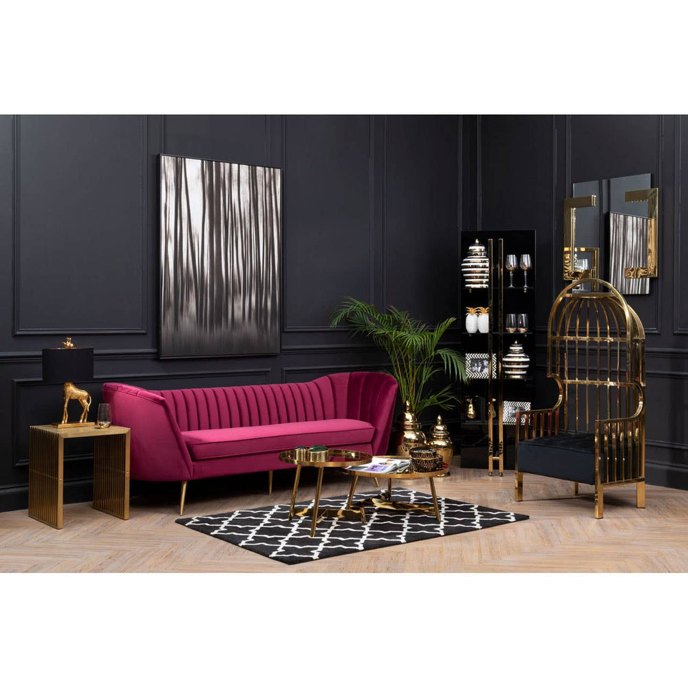 A vibrant wine velvet sofa with three seats is positioned against a dark wall, complemented by a stylish black and white patterned rug. A modern coffee table sits in front of the sofa, alongside a small side table with decorative items. A tall bookshelf filled with various decor pieces and a large framed artwork are visible in the background, along with a potted plant adding a touch of greenery to the scene