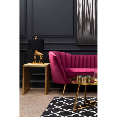 A plush wine velvet sofa with a curved backrest and vertical tufting, positioned against a dark wall. Next to the sofa, there is a small gold side table with a decorative lamp featuring a unique animal-shaped base. A patterned black and white rug is visible on the floor, adding contrast to the scene