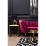 A plush wine velvet sofa with a curved backrest and vertical tufting, positioned against a dark wall. Next to the sofa, there is a small gold side table with a decorative lamp featuring a unique animal-shaped base. A patterned black and white rug is visible on the floor, adding contrast to the scene