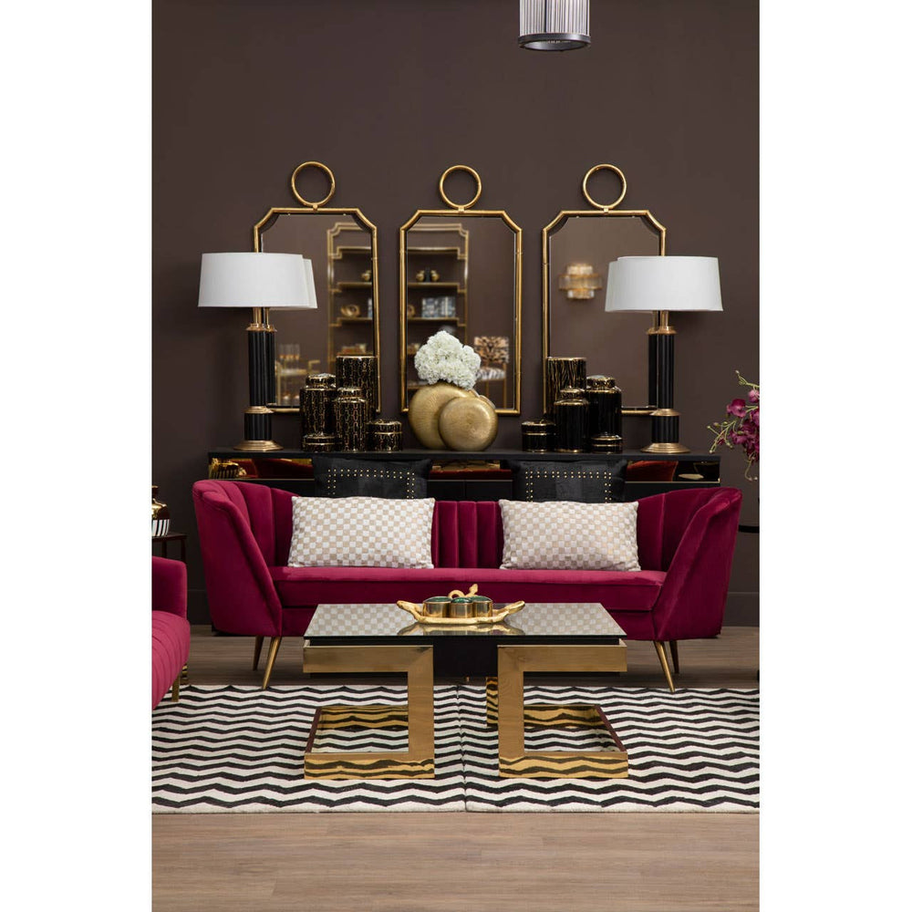 A luxurious three-seat sofa upholstered in rich wine-colored velvet, featuring elegant curved arms and plush cushions. The sofa is positioned in a stylish living room setting with a dark brown wall, complemented by a decorative gold-framed mirror and matching lamps on either side. A modern coffee table with a gold base sits in front of the sofa, resting on a patterned area rug