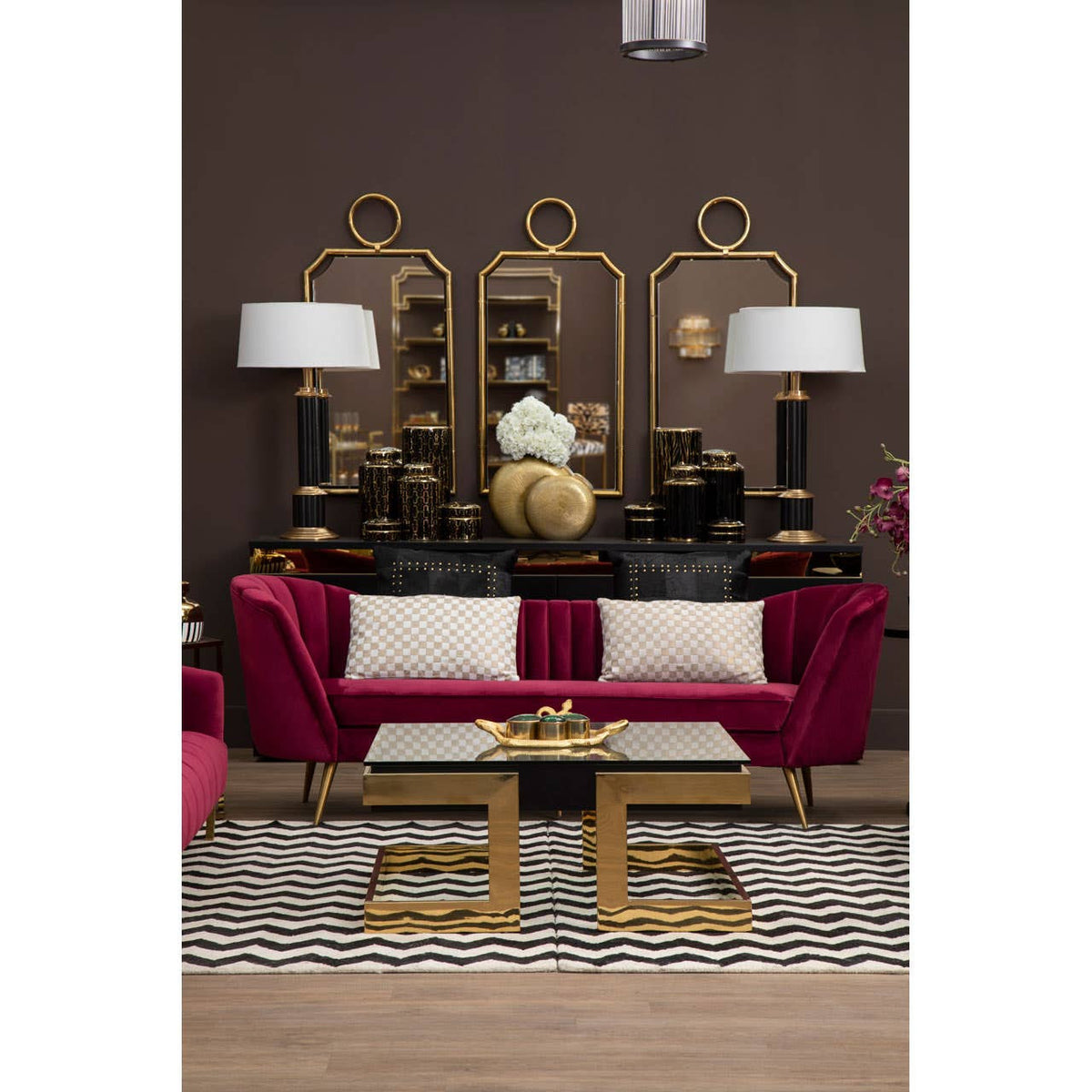 A luxurious three-seat sofa upholstered in rich wine-colored velvet, featuring elegant curved arms and plush cushions. The sofa is positioned in a stylish living room setting with a dark brown wall, complemented by a decorative gold-framed mirror and matching lamps on either side. A modern coffee table with a gold base sits in front of the sofa, resting on a patterned area rug