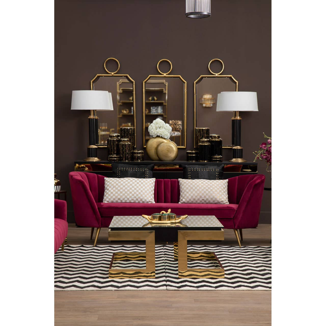 A luxurious three-seat sofa upholstered in rich wine-colored velvet, featuring elegant curved arms and plush cushions. The sofa is positioned in a stylish living room setting with a dark brown wall, complemented by a decorative gold-framed mirror and matching lamps on either side. A modern coffee table with a gold base sits in front of the sofa, resting on a patterned area rug