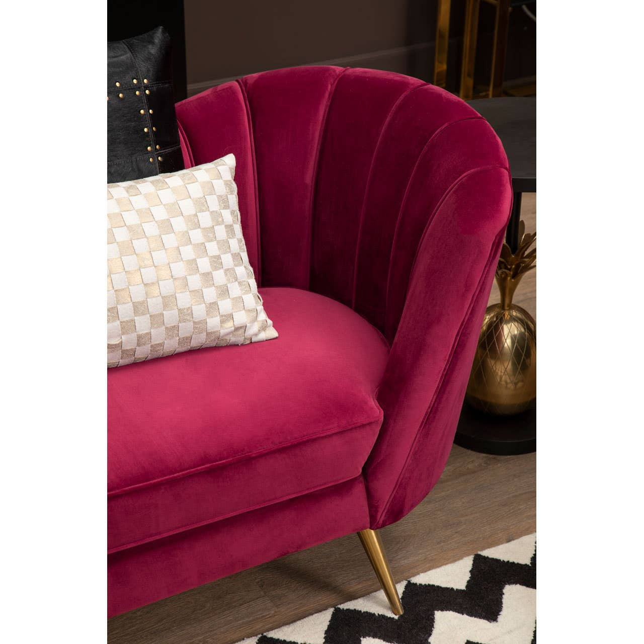 A luxurious wine-colored velvet sofa featuring a curved backrest with vertical pleats, complemented by gold metal legs. A decorative pillow with a geometric pattern rests on the seat, enhancing the elegant design. The sofa is positioned on a patterned black and white rug, with a blurred background showcasing a stylish interior setting