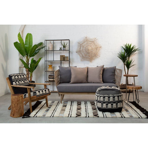 A black and white shaggy pouffe sits on a patterned rug in a modern living room setting. The room features a gray sofa with cushions, wooden furniture, and decorative plants, creating a cozy and stylish atmosphere