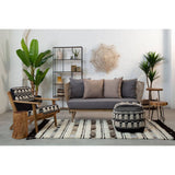 A black and white shaggy pouffe sits on a patterned rug in a modern living room setting. The room features a gray sofa with cushions, wooden furniture, and decorative plants, creating a cozy and stylish atmosphere