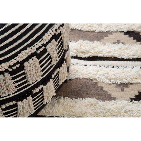 A black and white shaggy pouffe featuring a geometric pattern, placed on a textured, light-colored rug with subtle designs. The pouffe has a round shape and a soft, fluffy texture, contrasting with the smooth surface of the rug underneath